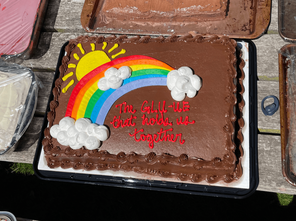 A chocolate cake with a frosting rainbow in the top left corner. The red icing text on the right side of the cake says "the GLU-UE that holds us together."