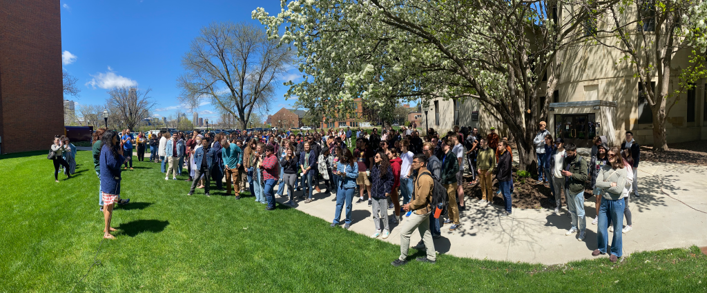A crowd gathered on a lawn on the East Bank campus to celebrate May Day with GLU-UE. Attendees are listening to speeches. 