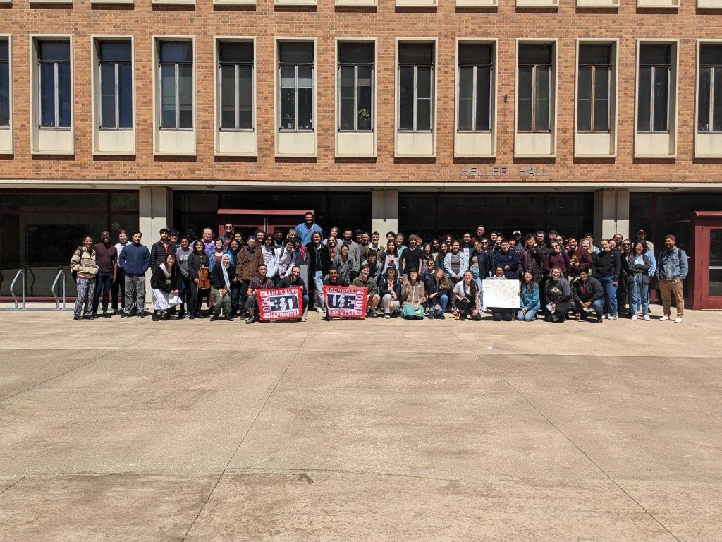 A group photo of the union break attendees on the West Bank campus. The UE flag and a poster board is held up in the front row.