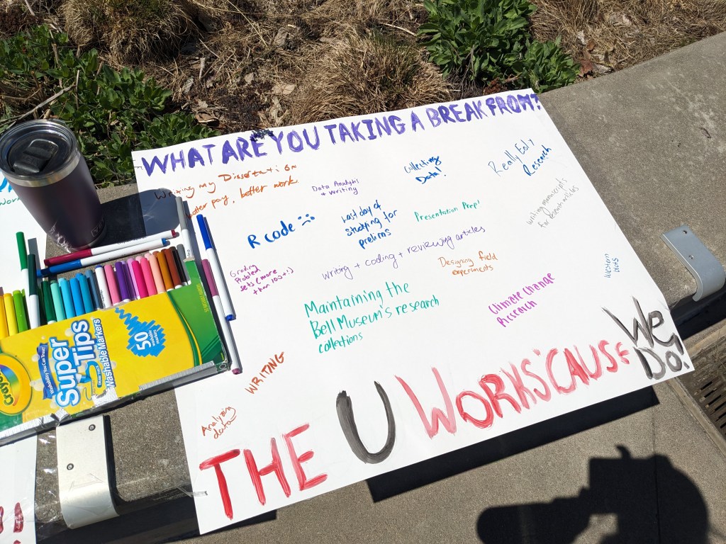 A poster board that says "What are you taking a break from?" at the top in purple text. Some written blurbs say "R code" with a frown-y face, "Maintaining the Bell Museum's research collections", "climate change research", and "writing." "The U Works 'Cause We Do!" is painted at the bottom in red and black paint.